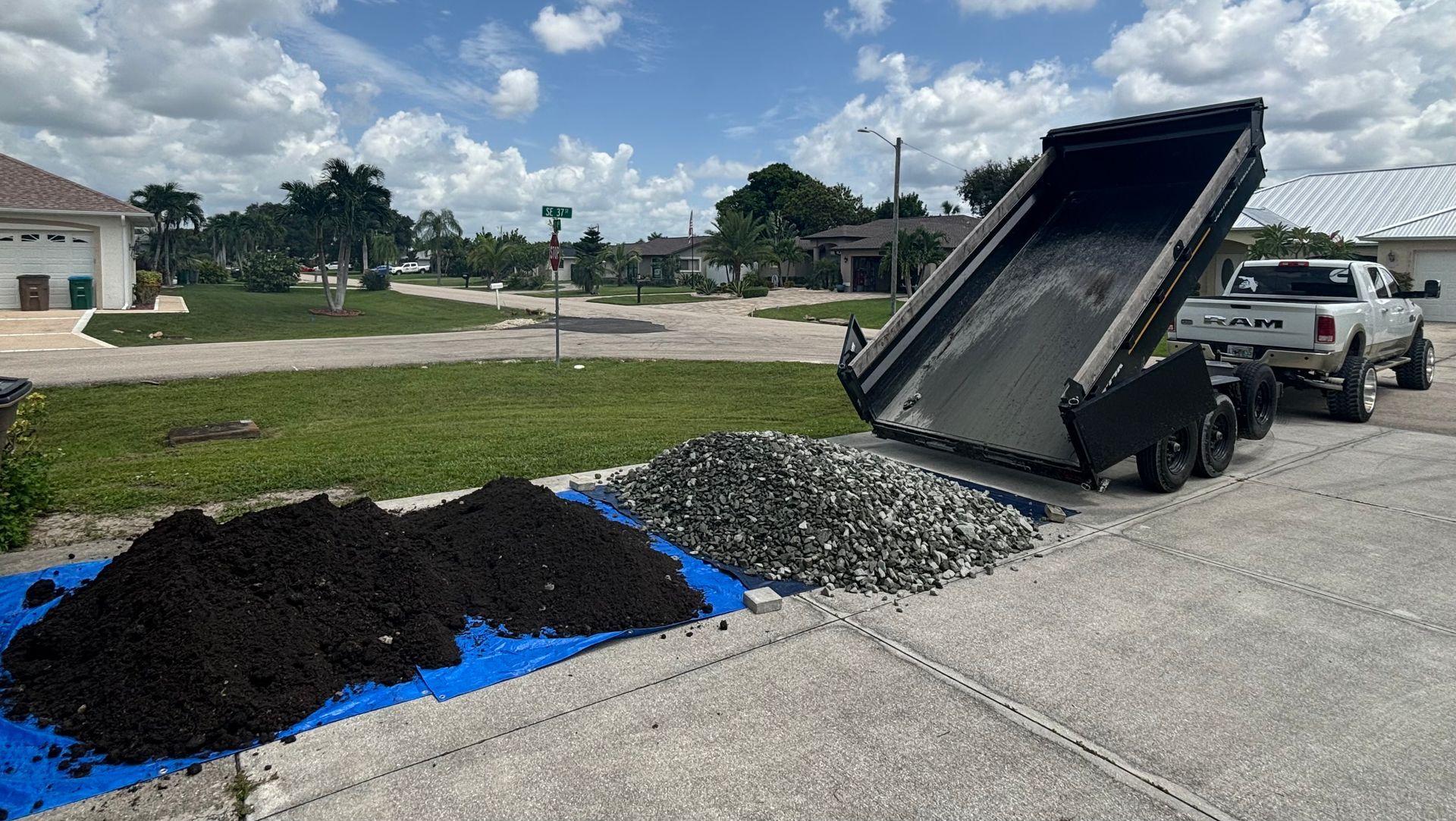 A truck dumping gravel and dark mulch onto a driveway, with piles on a blue tarp.