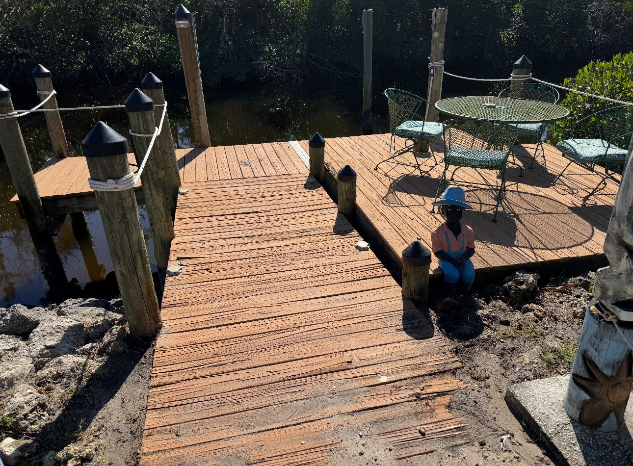Wooden dock leading to a waterside deck with a table, chairs, and a statue; sunny day.