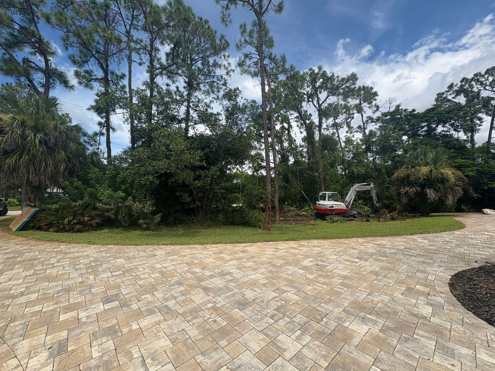 Construction site: An excavator clearing brush behind a brick-paved driveway, with trees in the background.