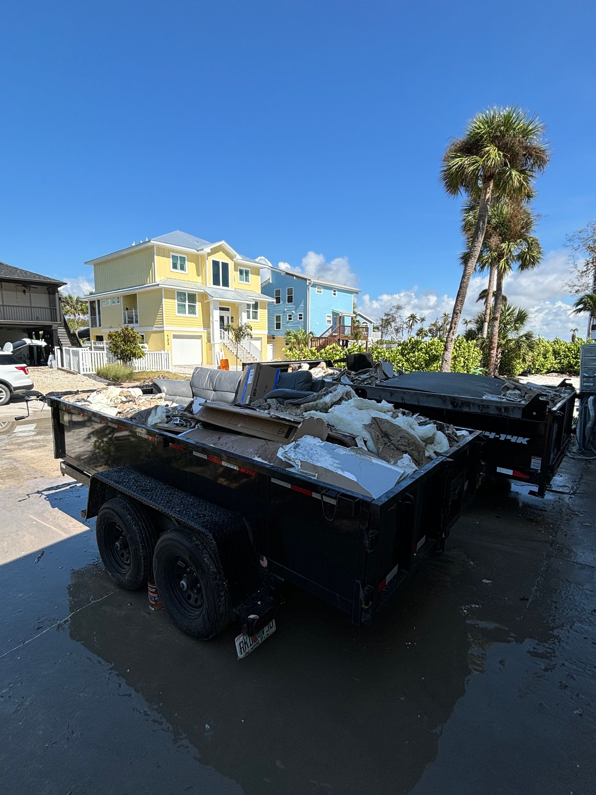 Two black trailers loaded with debris sit in a flooded area, with two-story yellow and blue houses in the background.