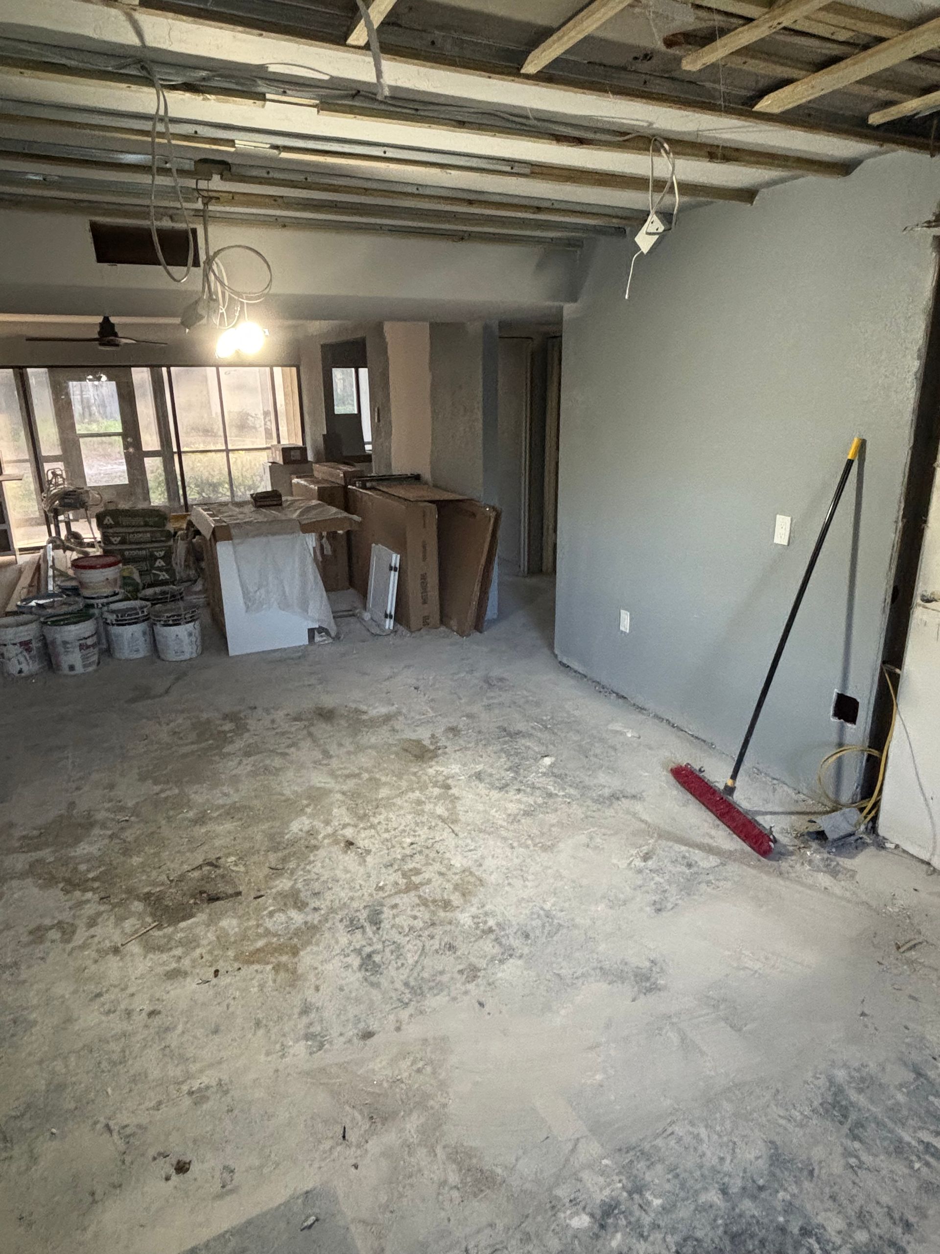 Interior construction site, concrete floor, gray walls, unfinished ceiling, building materials, red broom leaning against a wall.