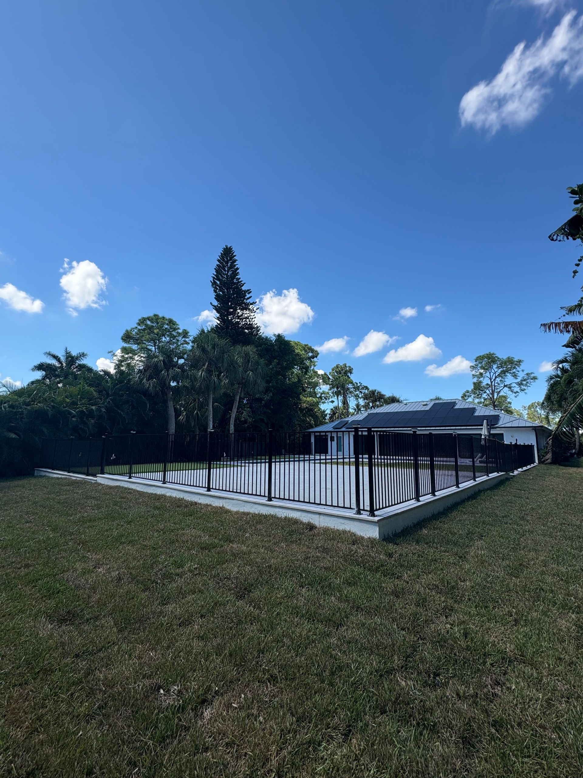 Pool with black fence, gray deck, blue sky with clouds, green trees and grass.
