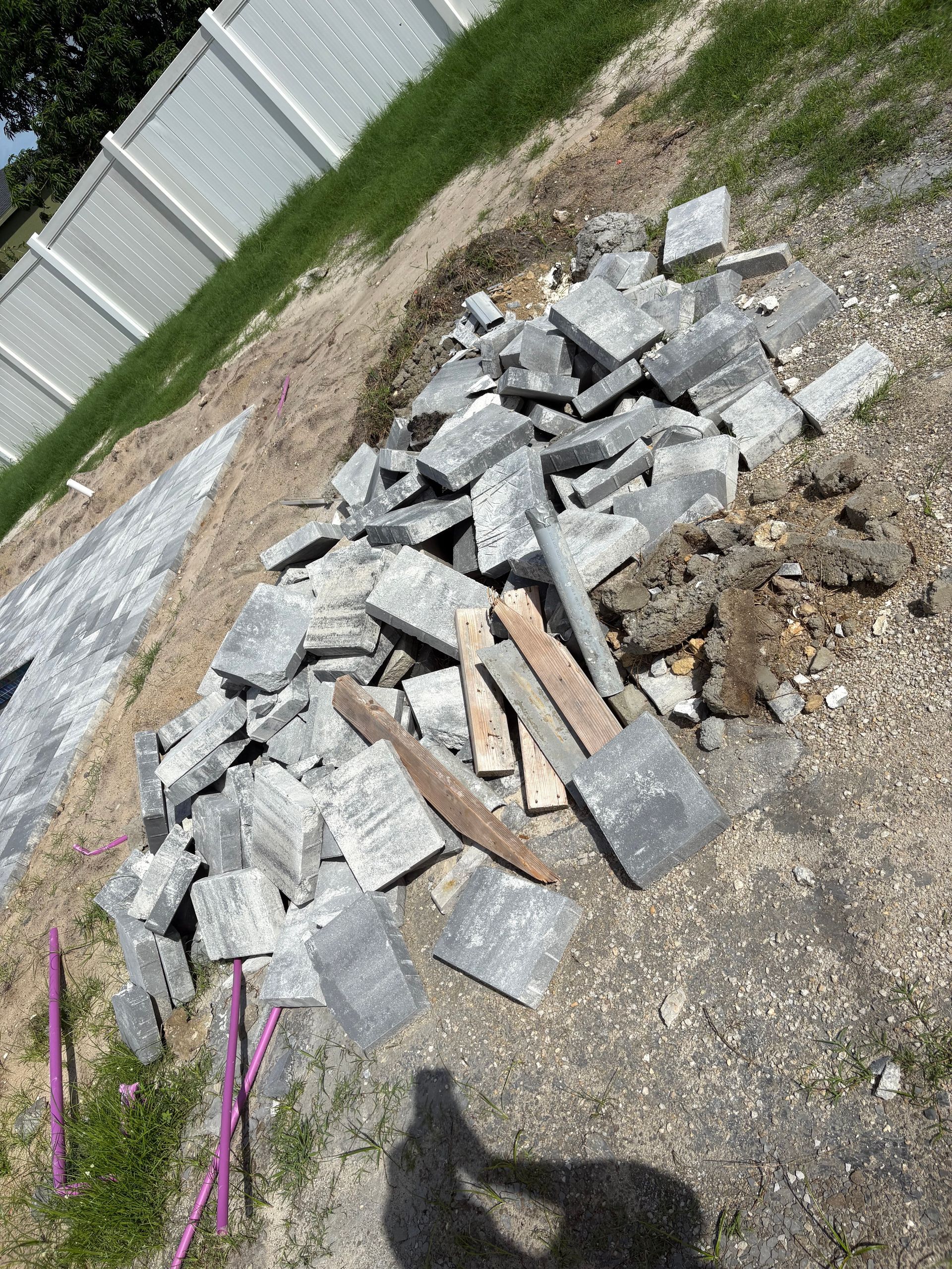 Pile of broken gray paving stones, wood, and dirt on a gravel surface next to a white fence and grass.