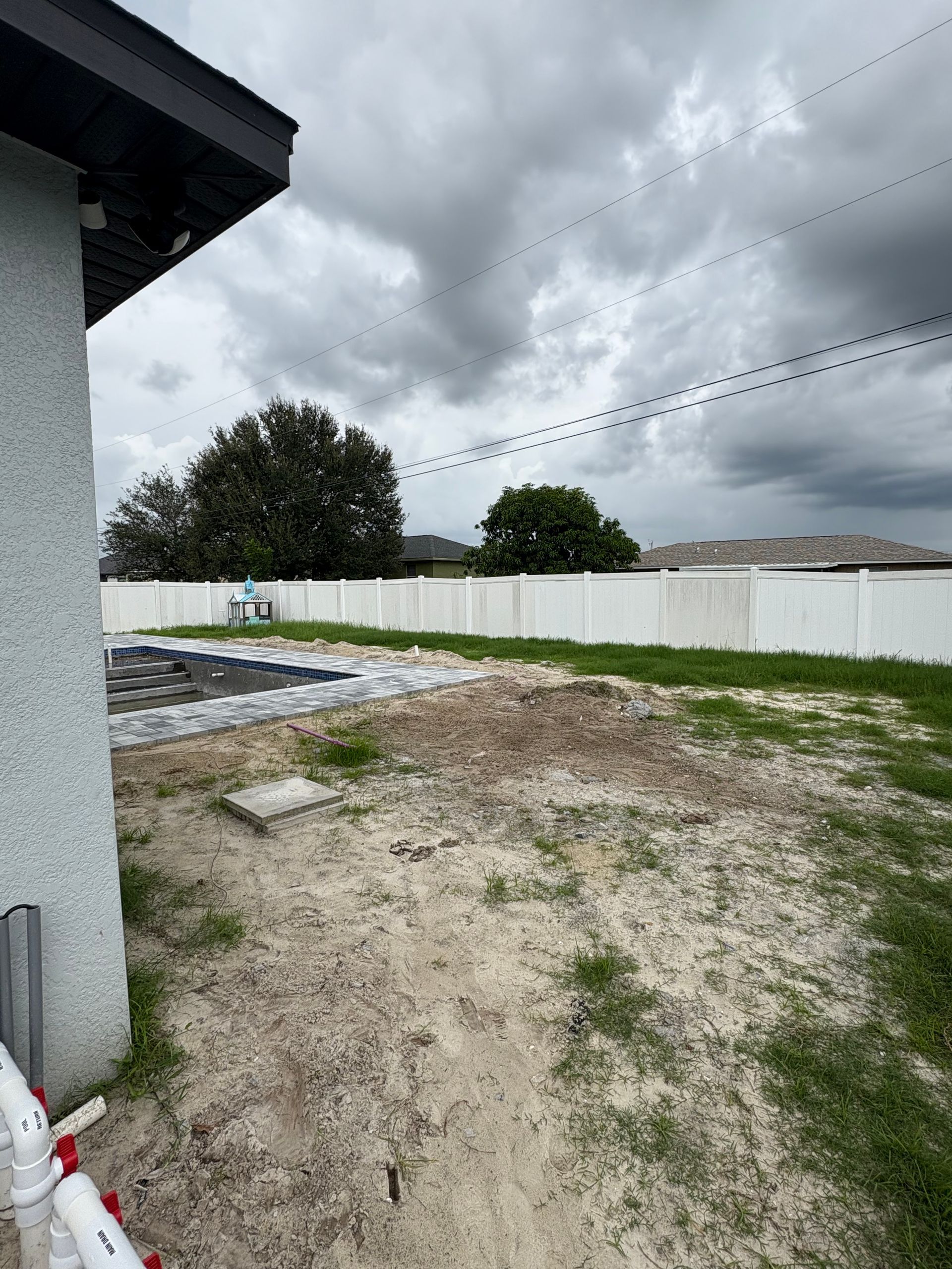 Backyard with white fence, pool, dirt patch, cloudy sky.