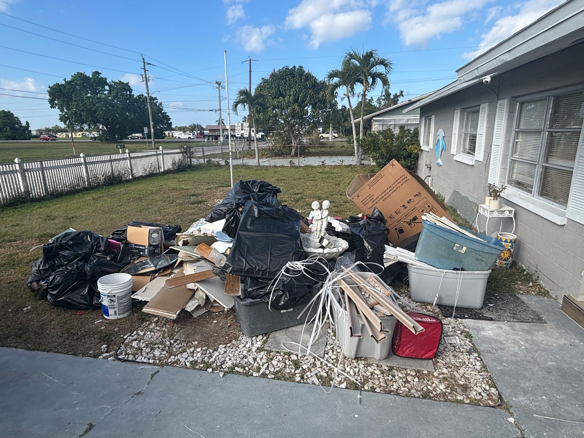 Pile of trash, including black bags, boxes, and debris, in front of a house on a sunny day.