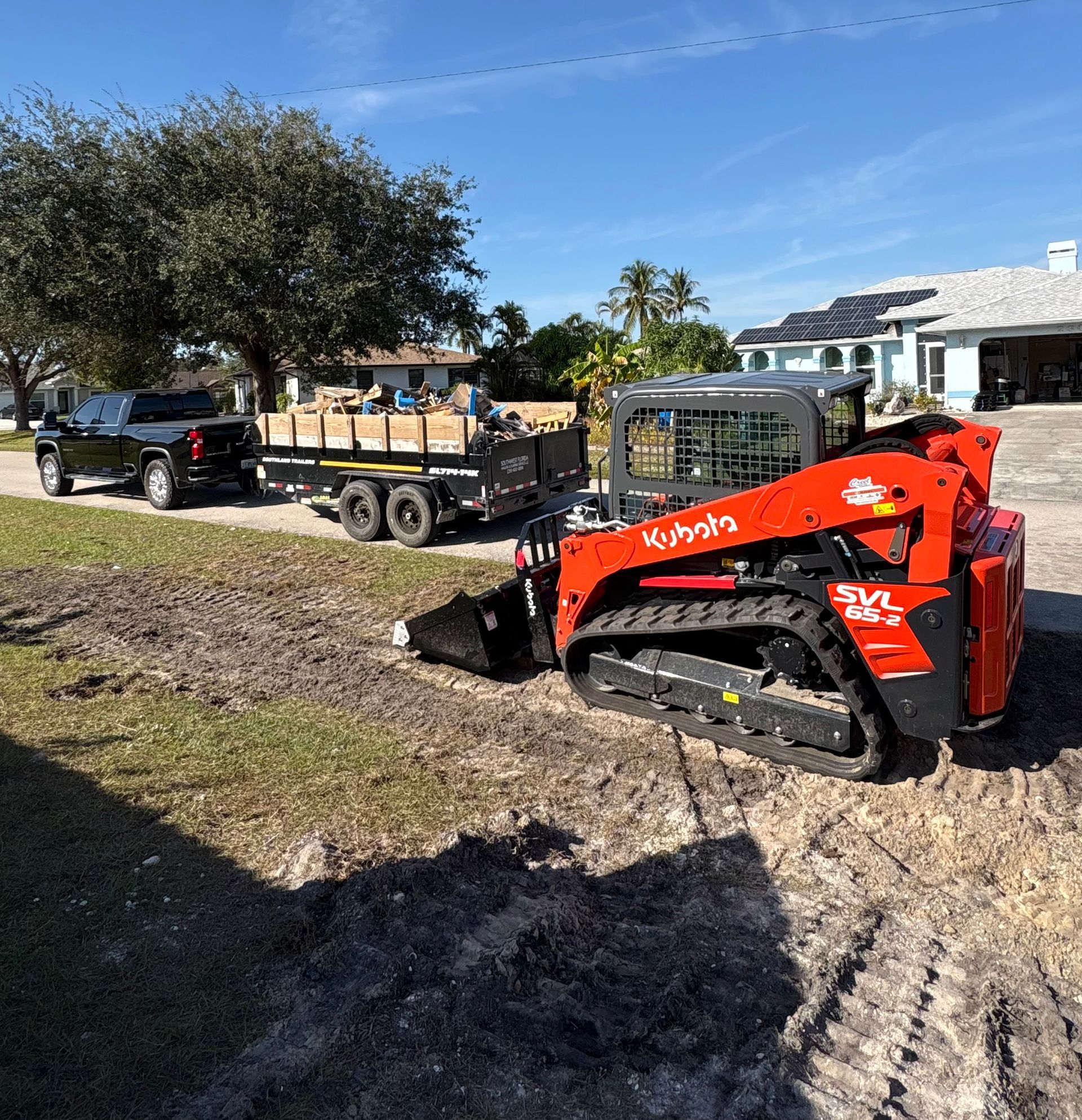 Orange Kubota skid steer on dirt near a truck and trailer loaded with materials on a sunny day.