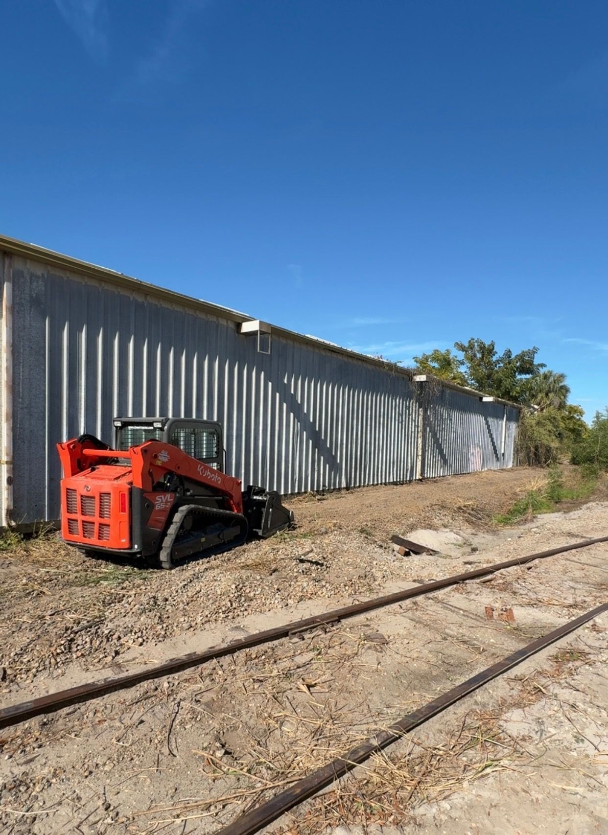 Orange skid steer loader next to a corrugated metal building and railroad tracks under a blue sky.