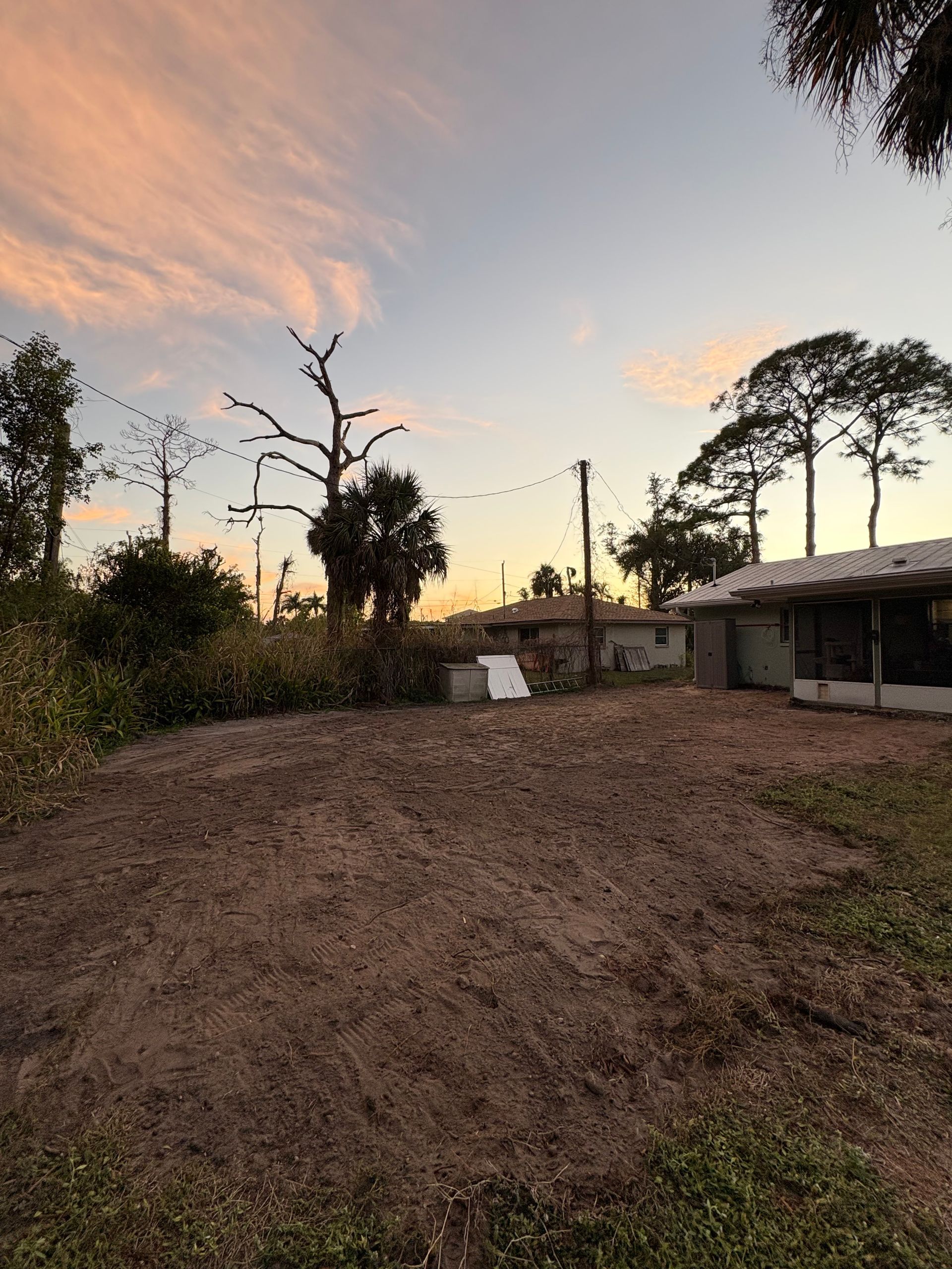 Dirt field with greenery, trees, and small building under a sunset sky.