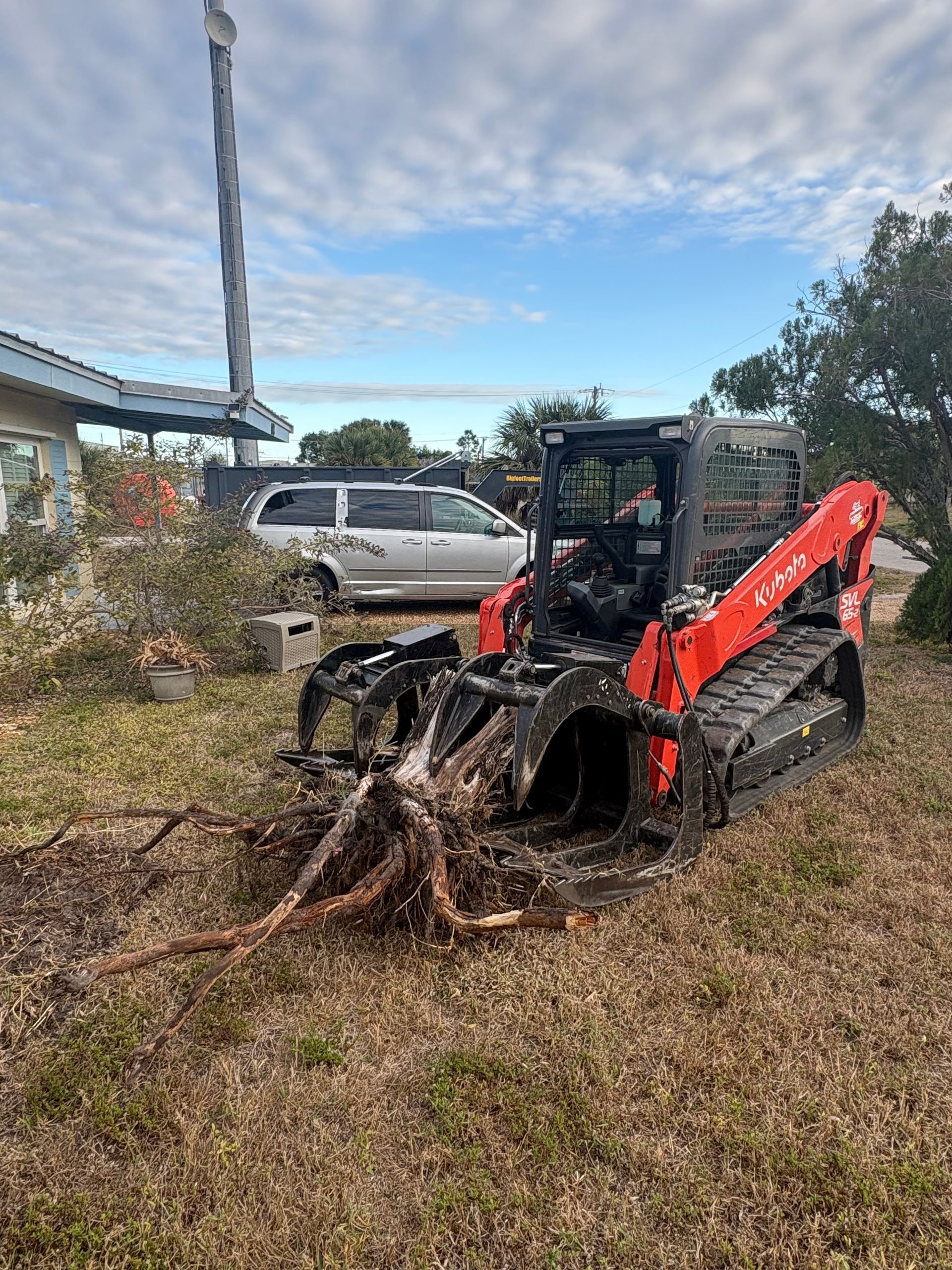Burnt orange skid steer on tracks in a yard, charred engine area, roots in front, car and house in background.