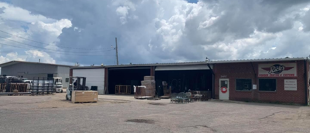 Wide gravel yard with a low industrial building, open bays, and a red office section under a cloudy sky