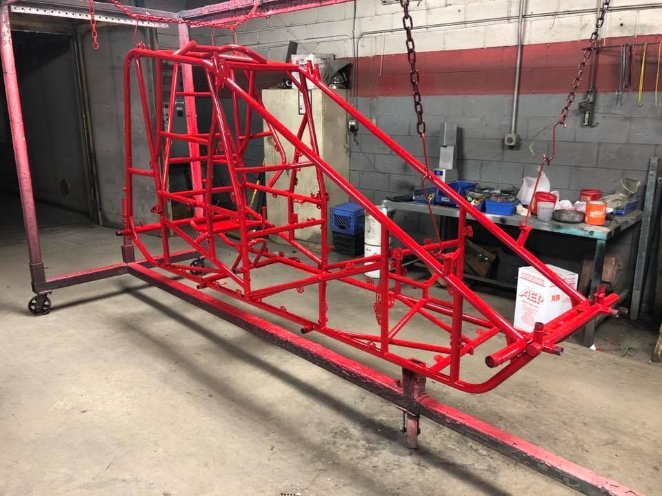 Red metal car chassis frame hanging in a workshop garage, with tools and parts in the background