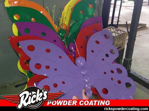 Colorful butterfly-shaped metal sign with purple, green, orange, and red wings outdoors, labeled Rick's Powder Coating