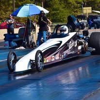 Dragster race car on a blue track, driver helmeted in cockpit, with crew and umbrella behind it