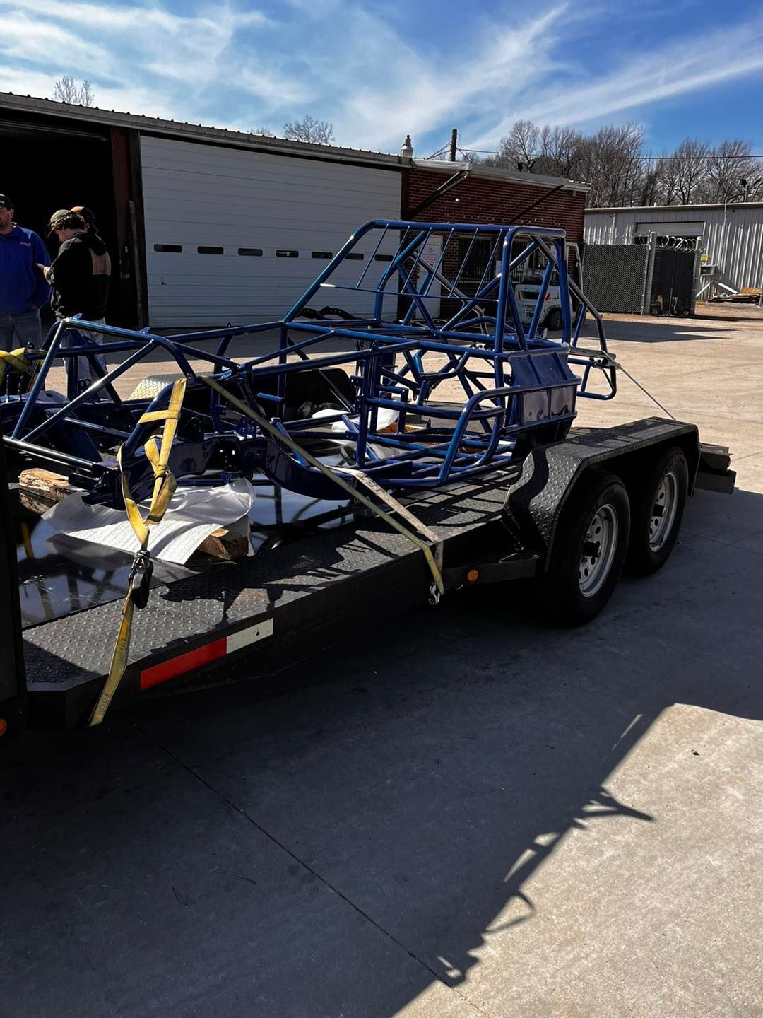 Blue race car frame strapped to a flatbed trailer in a driveway, with a garage in the background