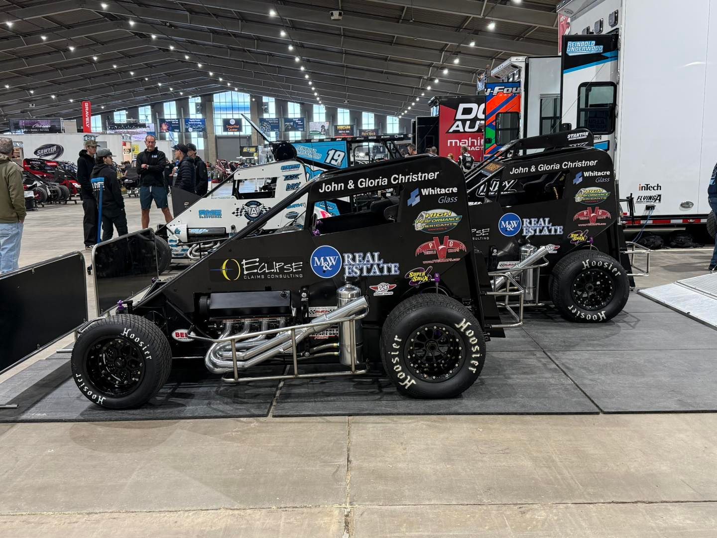 Black sprint car on display at an indoor motorsports show, with people and trailers in the background