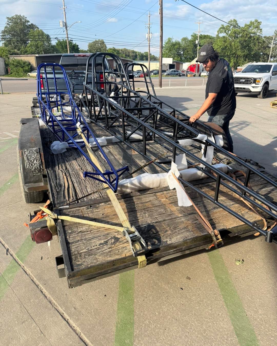 Man securing a large black metal framework on a trailer in a parking lot, with straps and blue tubing attached