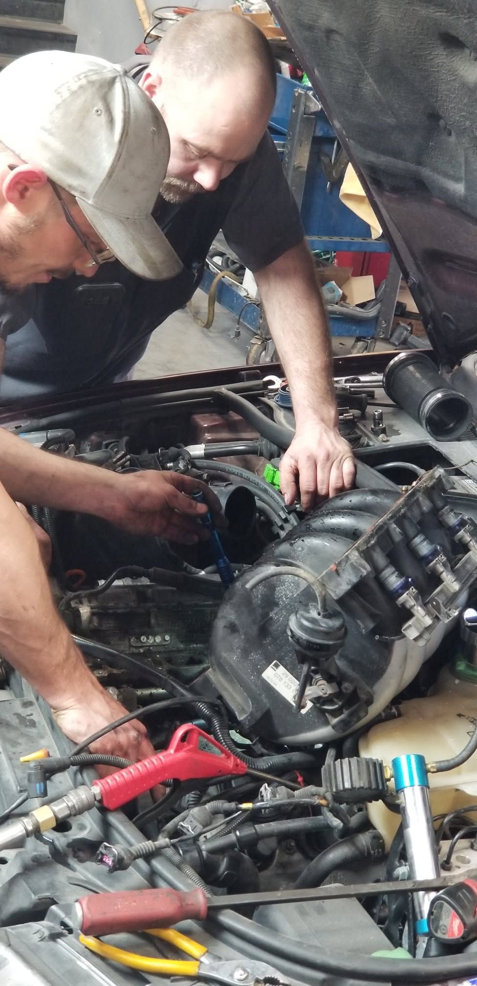 Two mechanics leaning over an open car engine in a garage, using tools for repair