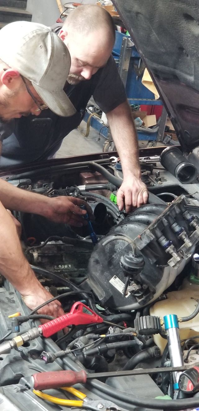 Two people working under a car hood, inspecting engine components with tools nearby.