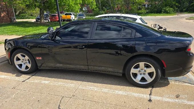 Black sedan parked at the curb on a sunny street, with trees and a white car in the background