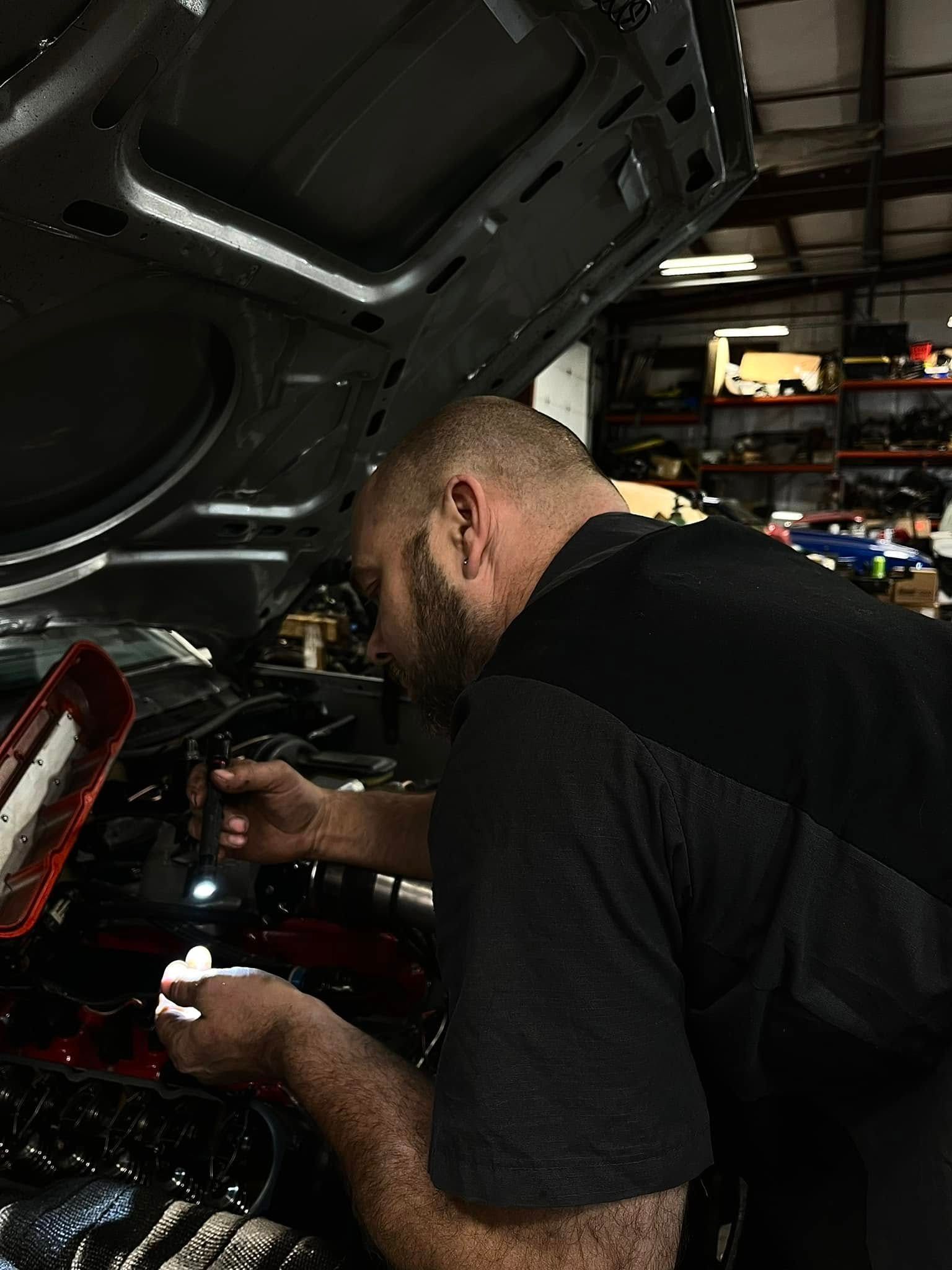 Mechanic working under an open car hood in a dim garage, using a tool near the engine.