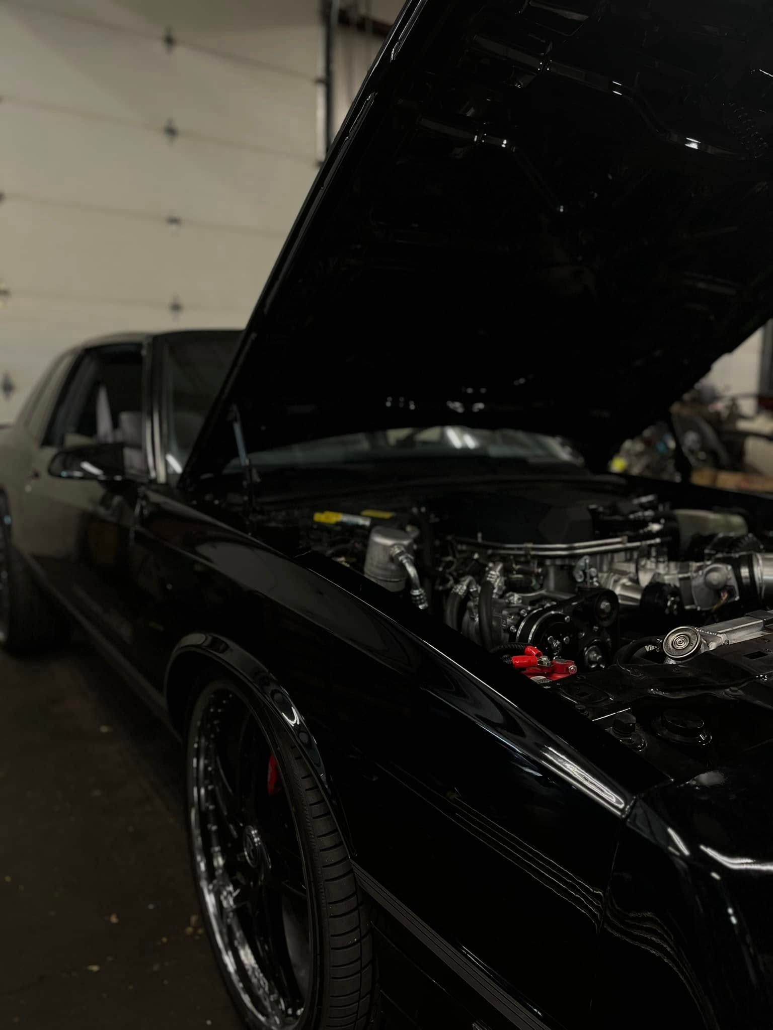 Black classic car with hood open, showing the engine in a dim garage.