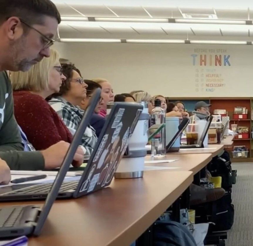 People seated at a long table with laptops, likely in a classroom or meeting. The word 