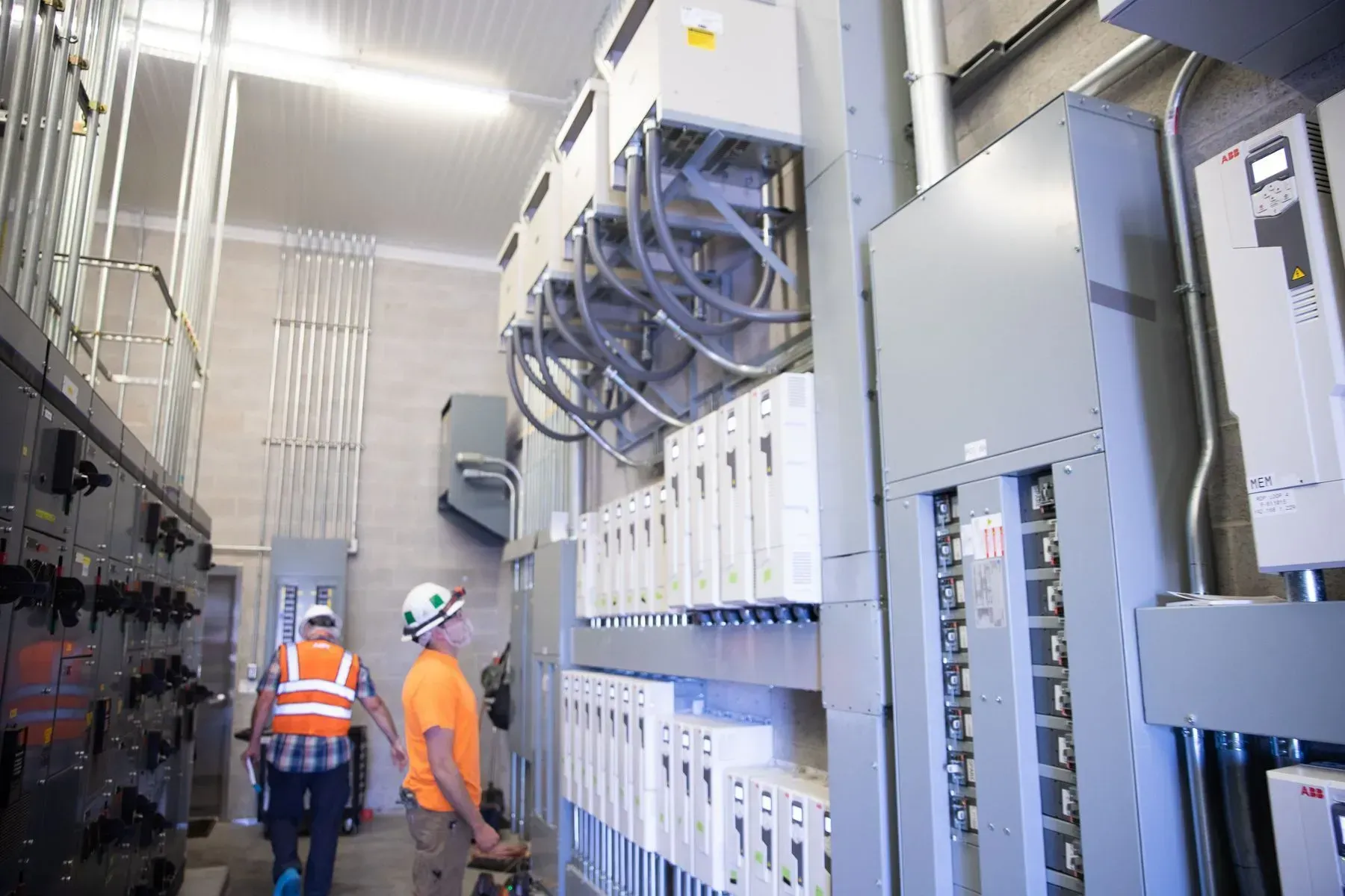 Two workers in safety gear inspect electrical panels and control equipment inside an industrial facility.