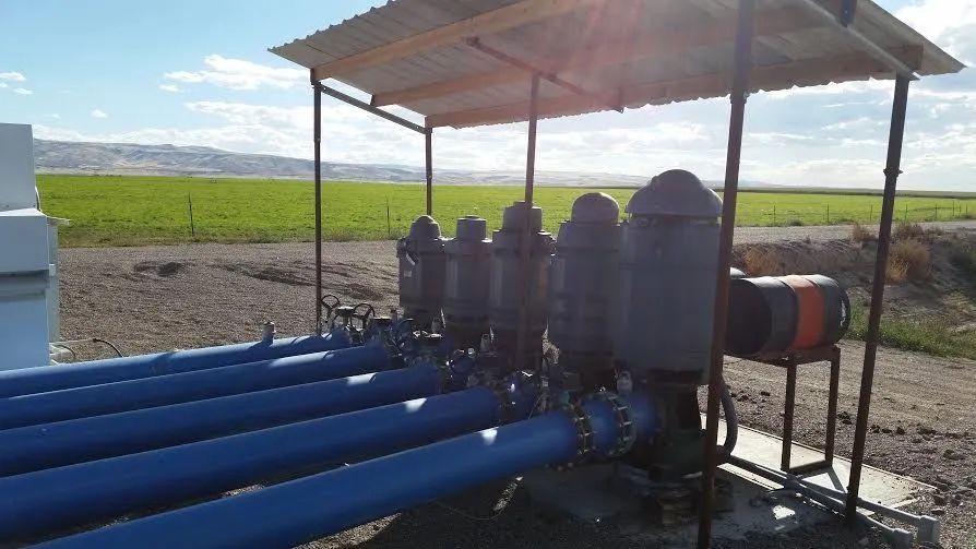A row of industrial agricultural filters under a roof, connected to blue pipes in an open field.