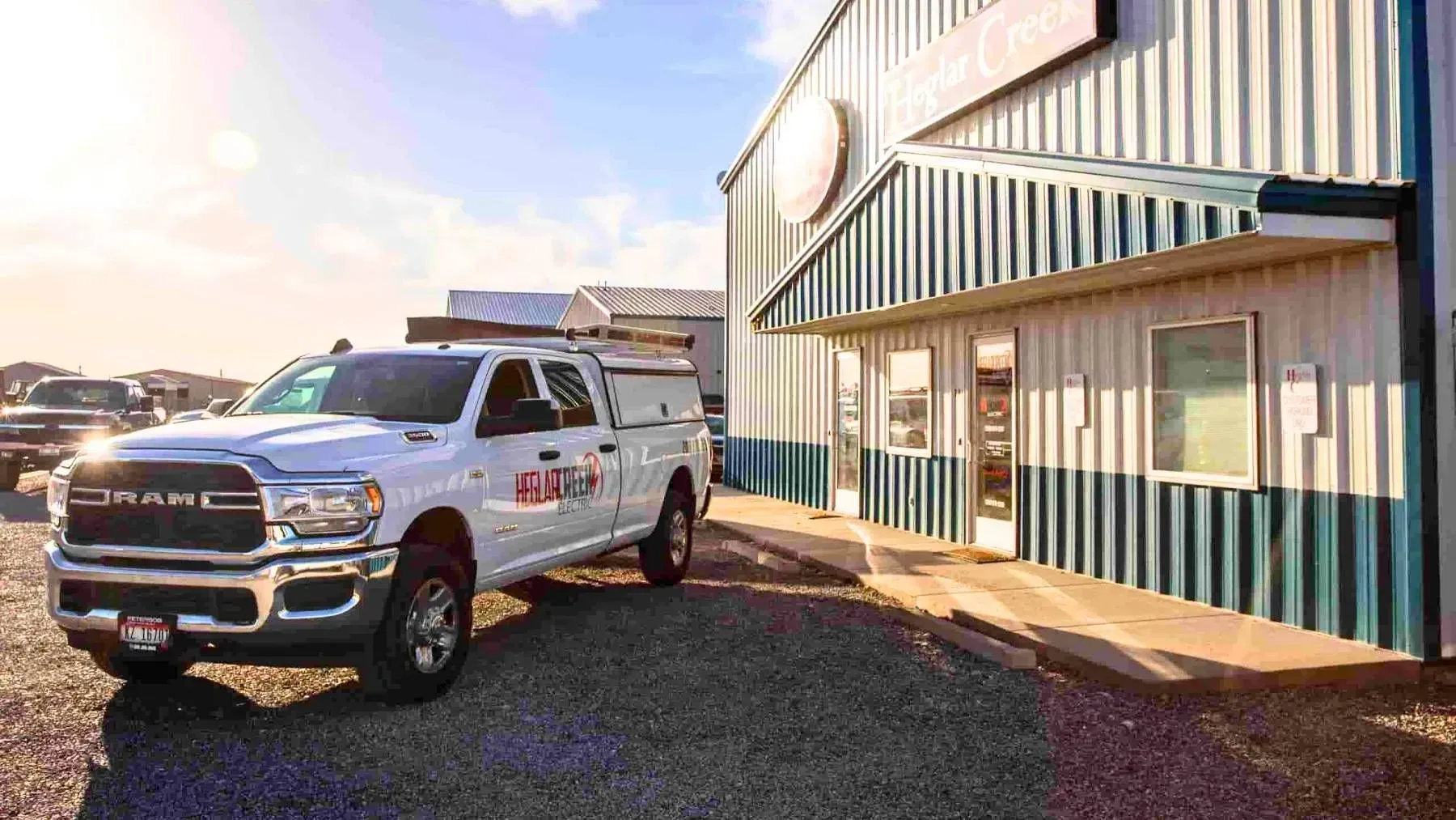 A white RAM service truck with a roof rack parked on a gravel lot in front of a modern metal building under a sunny sky.