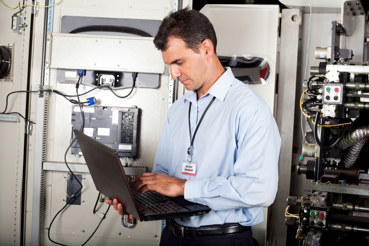 Technician in a light blue shirt using a laptop in an equipment room with electrical panels.