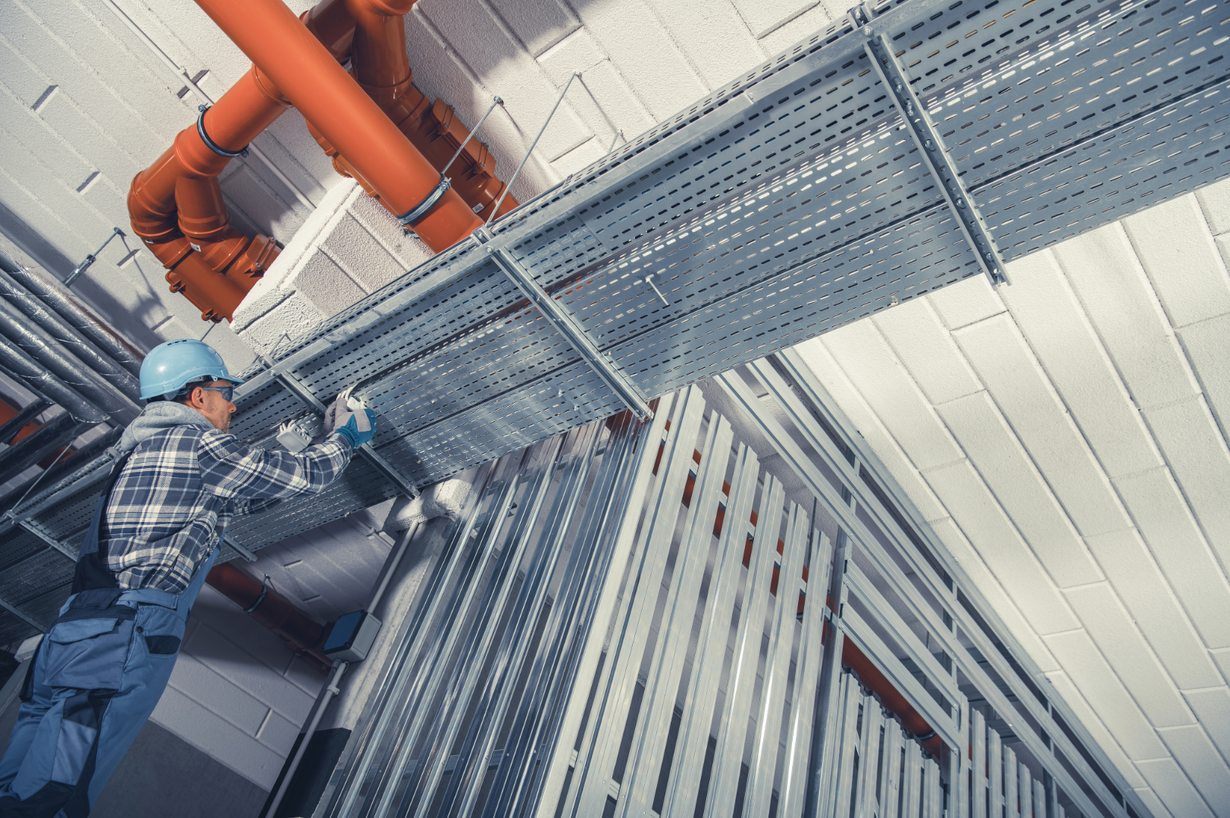 Worker in a blue hard hat inspects overhead pipes and metal ducting in an industrial building.