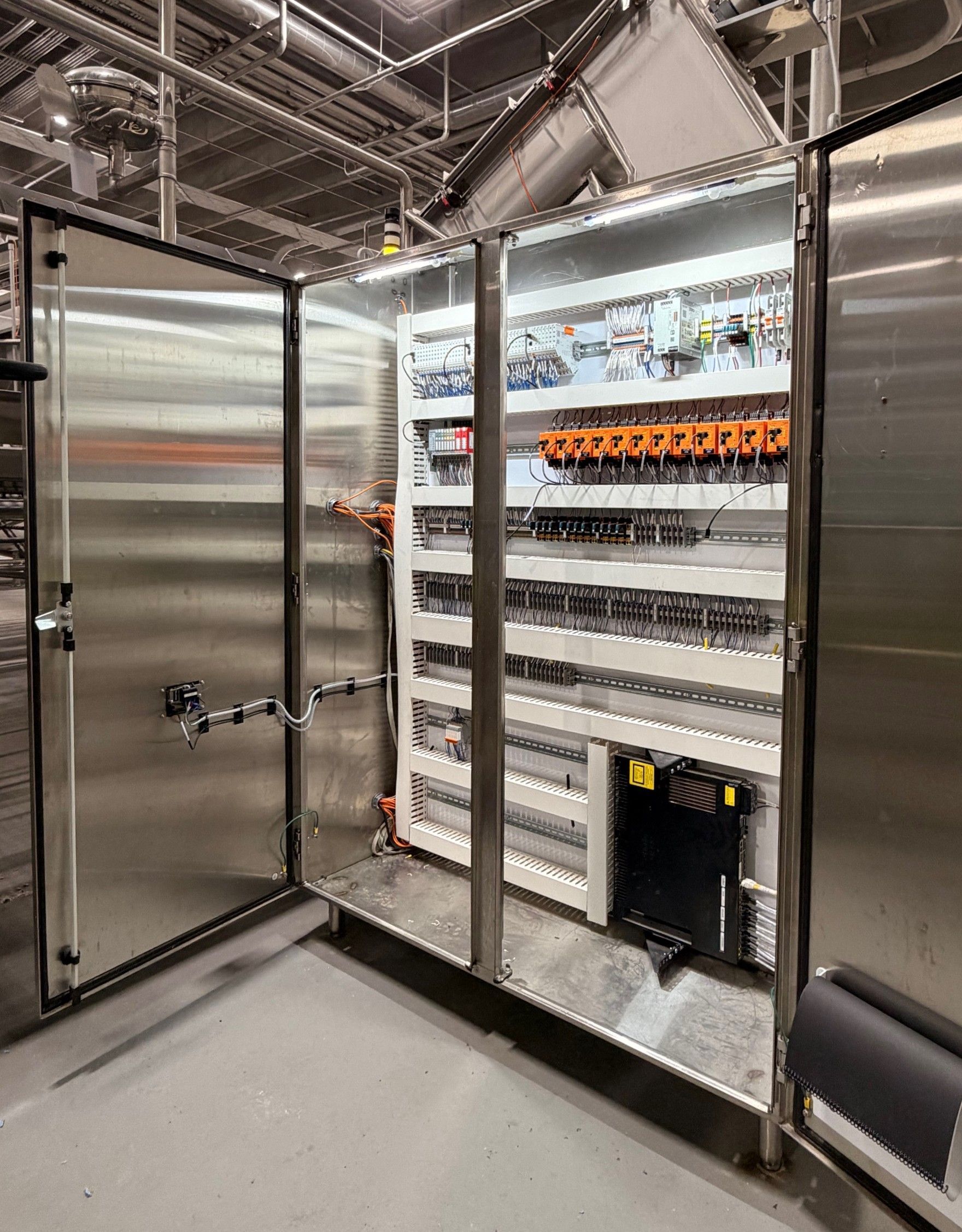 Open industrial control cabinet with wiring, circuit modules, and orange terminal blocks inside a factory.