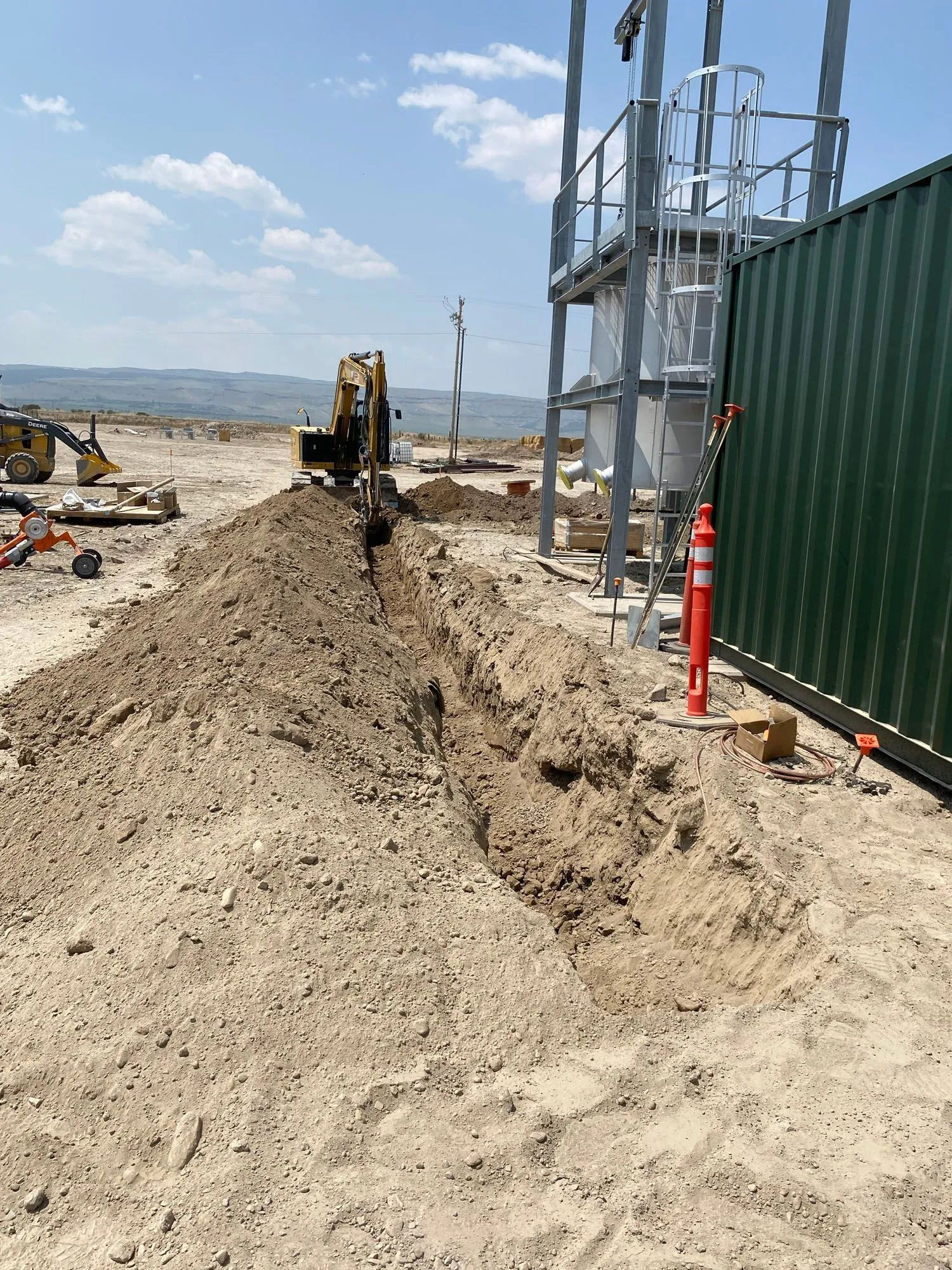 Construction site with dirt mound, excavator, and metal structure beside a green fence under a blue sky