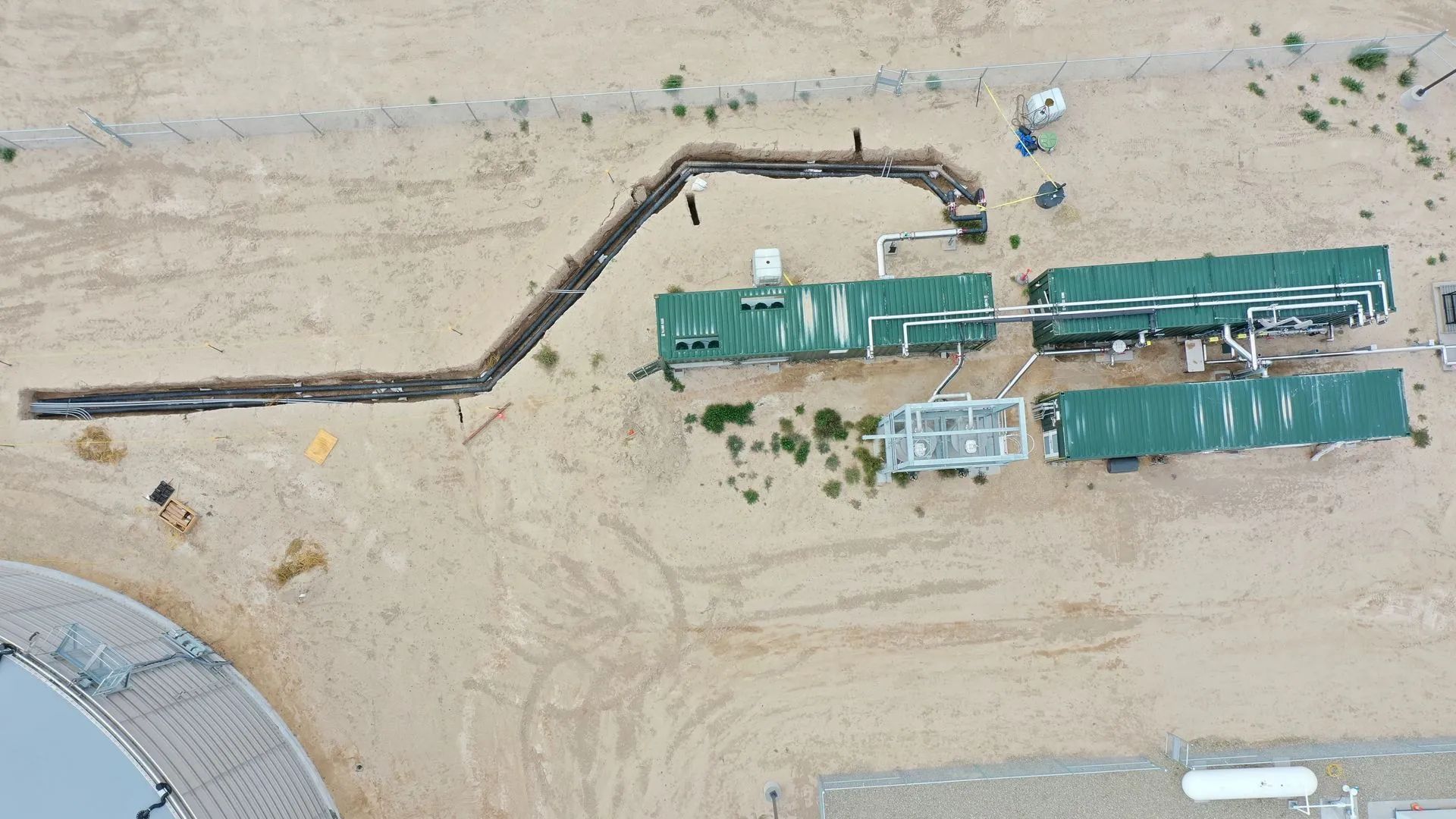 Aerial view of a sandy worksite with a long dark pipeline and several green temporary structures.