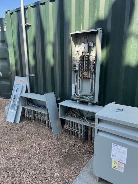 Electrical panels mounted on a green metal wall beside a gray utility box on gravel.