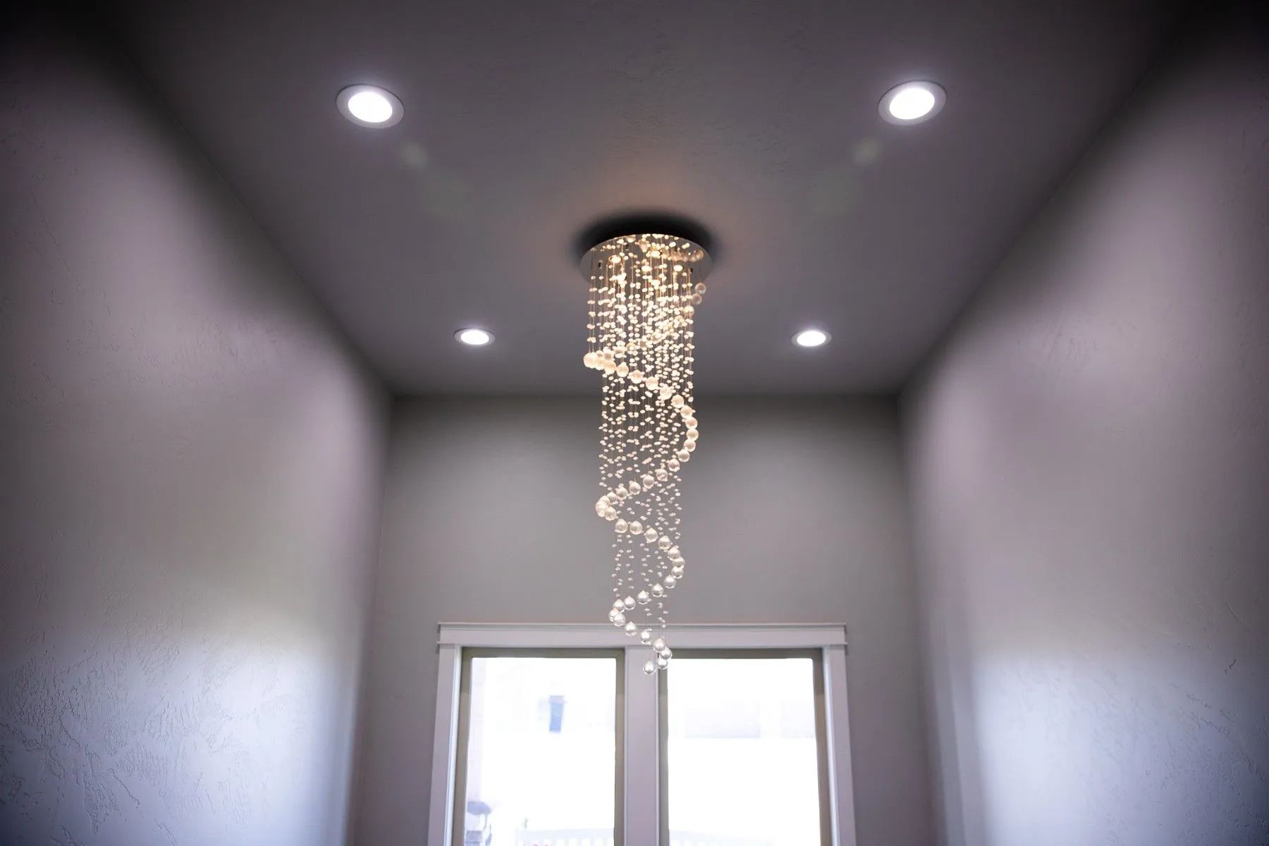 A spiral crystal chandelier hangs from a gray ceiling, illuminated by four recessed lights above a window.
