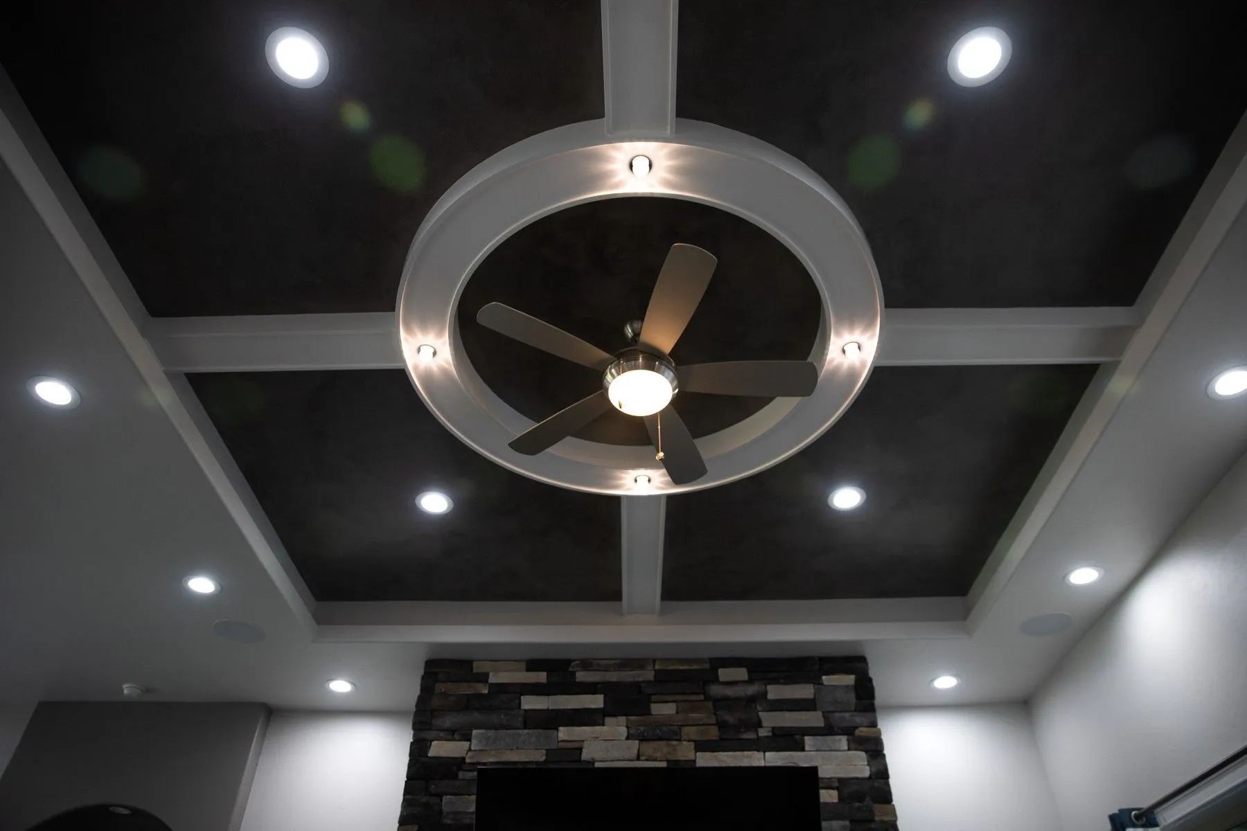 A modern room with a dark recessed ceiling, a circular feature holding a ceiling fan, and surrounding recessed lighting.