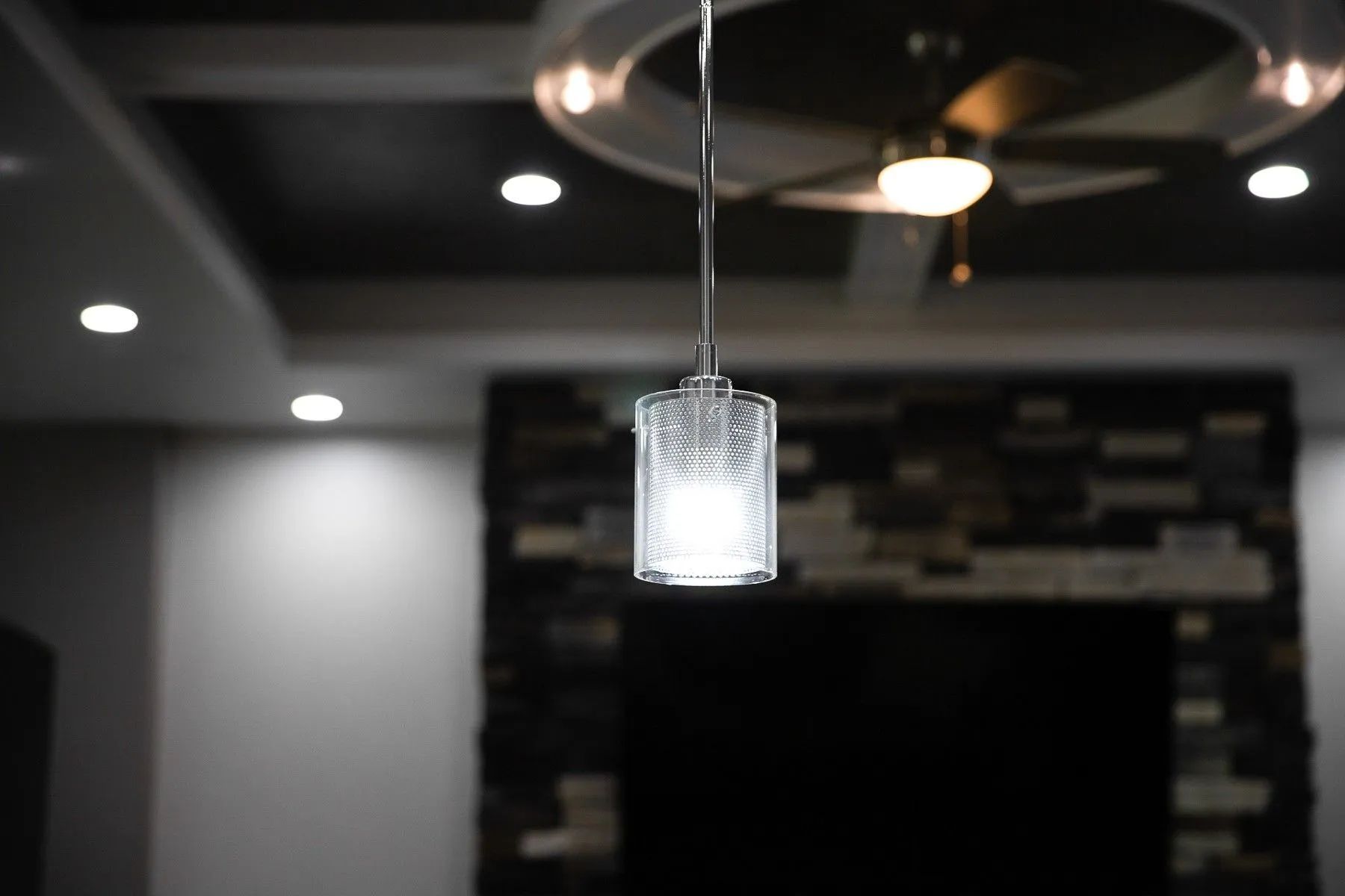 A textured glass pendant light glows brightly in the foreground, with a blurred ceiling fan and stone wall behind it.