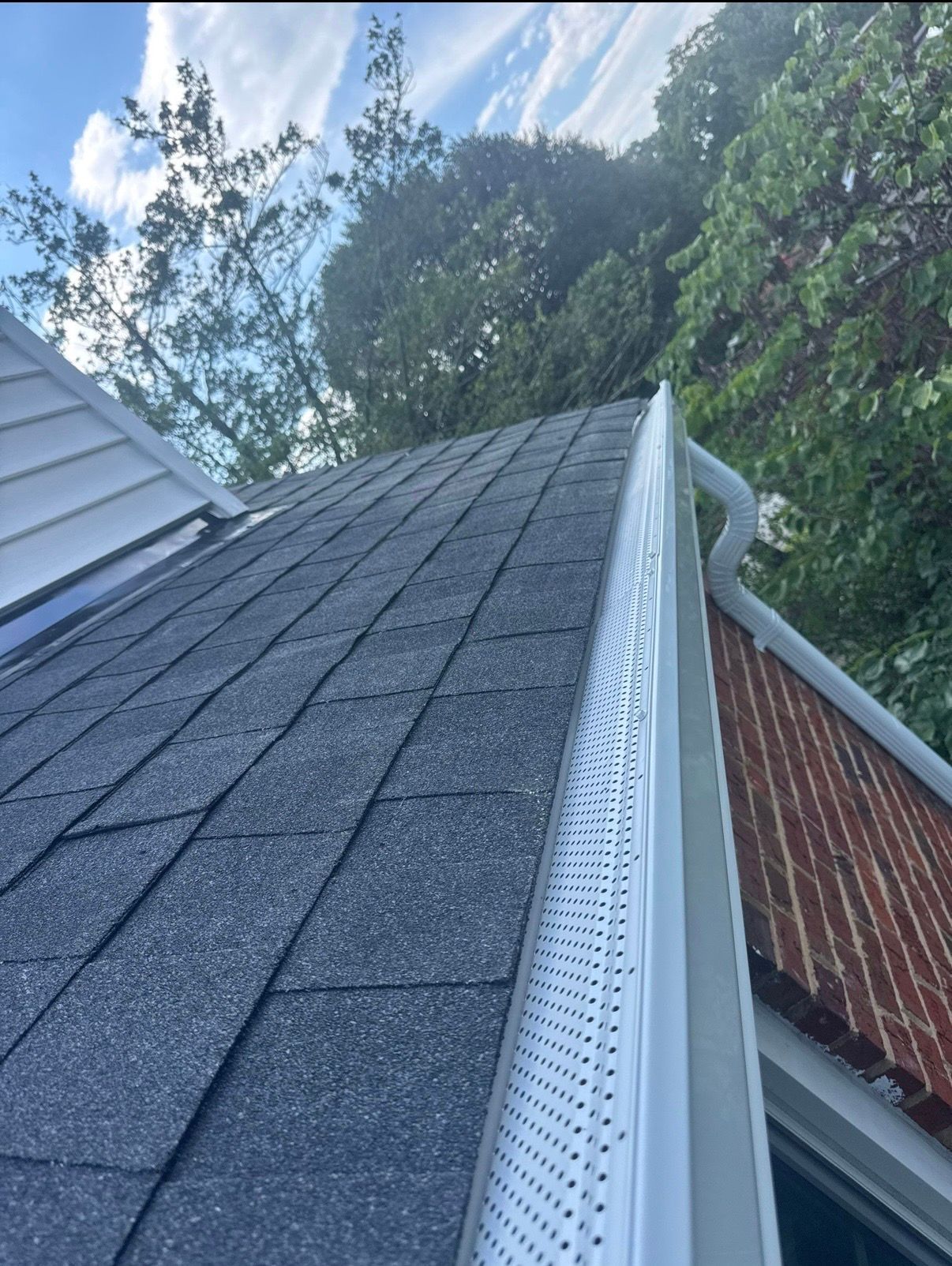 Roofline with dark shingles and white gutter against trees and blue sky