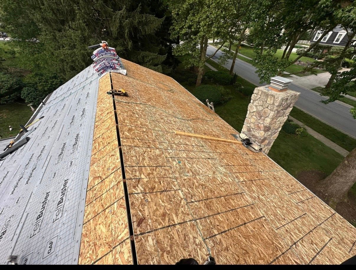 Roof under construction with plywood sheathing, a worker near the ridge, and a brick chimney on the right.