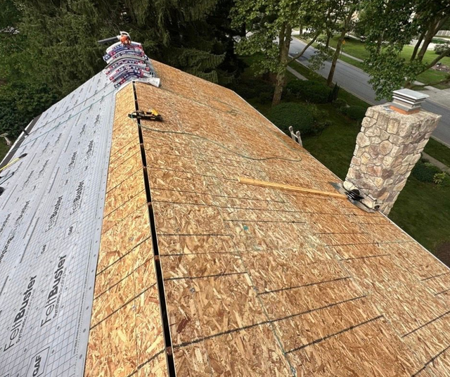 Roof under repair with plywood sheathing exposed and a worker kneeling near the ridge