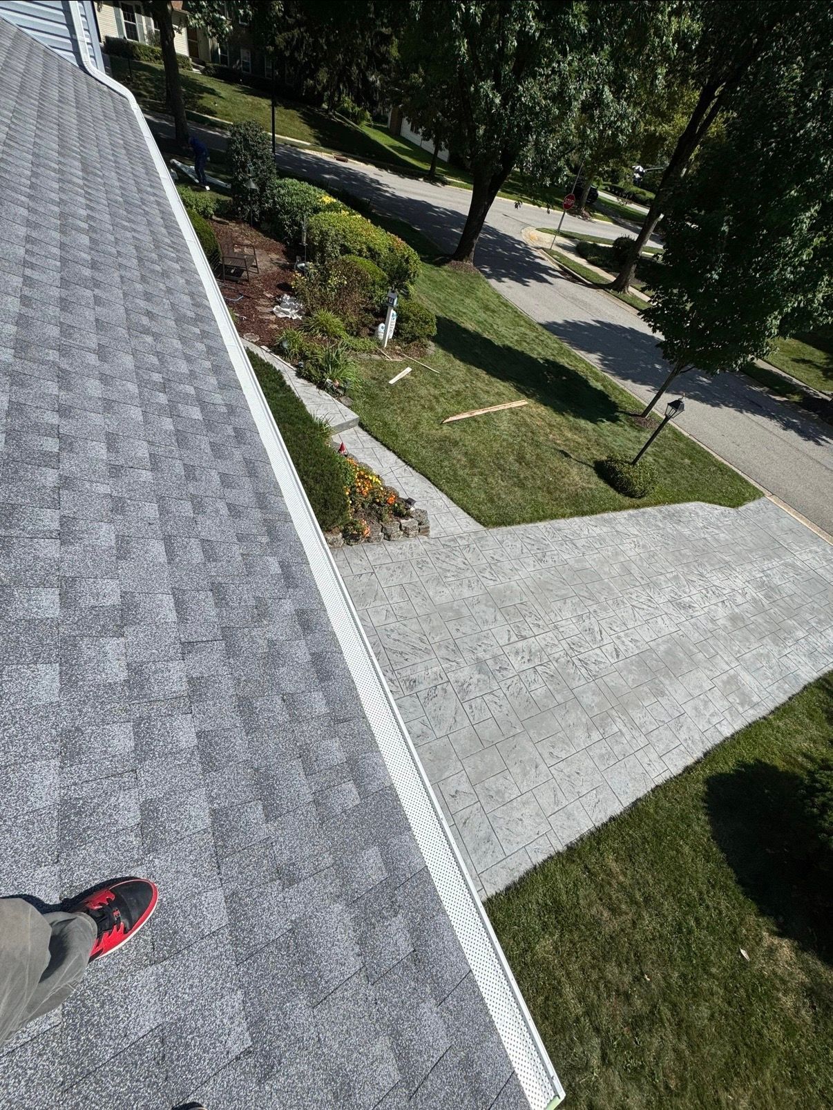 View from a roof edge onto a concrete walkway, lawn, and trees below, with a shoe visible at bottom left