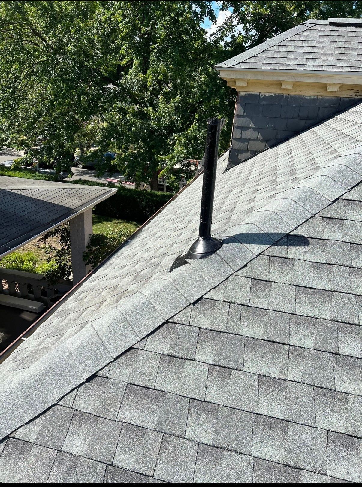 Steep gray shingle roof with a black vent pipe, trees and a covered patio in the background