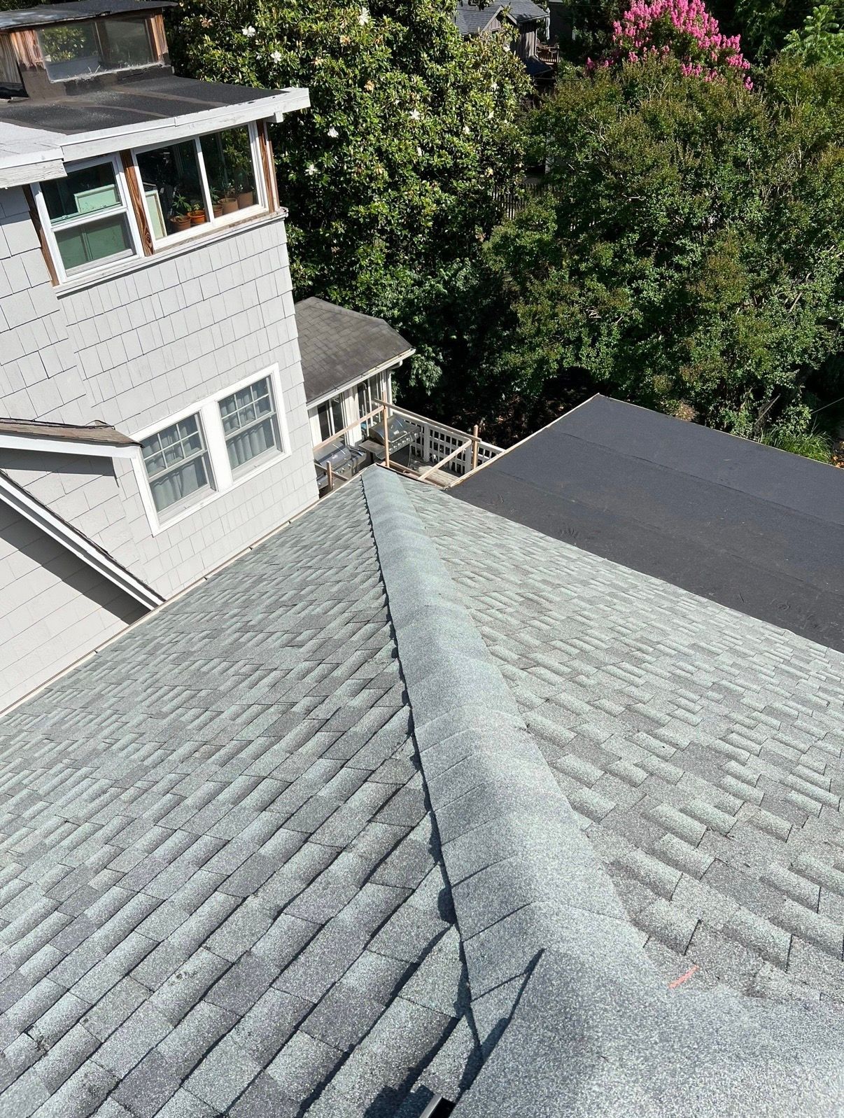 View from a gray shingled roof beside a house and trees, showing a roof valley and dark roof section.