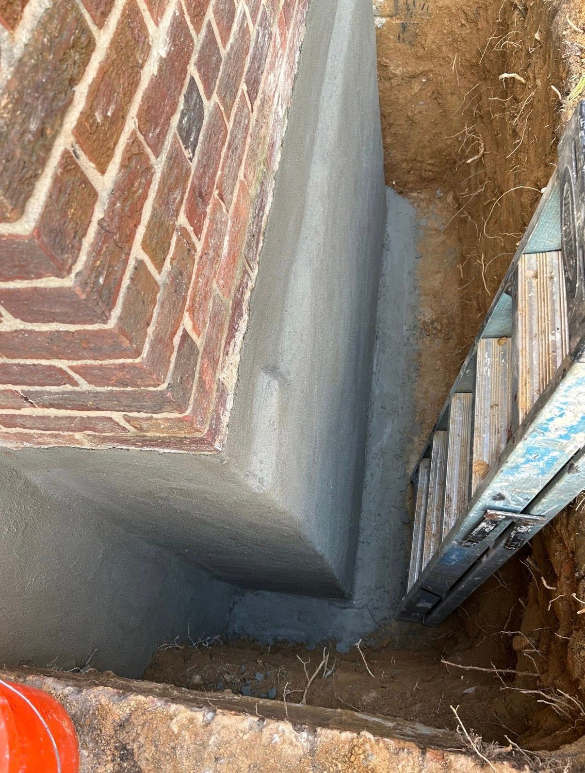 Narrow excavation beside brick foundation wall, with exposed soil and a metal ladder leaning in the trench.