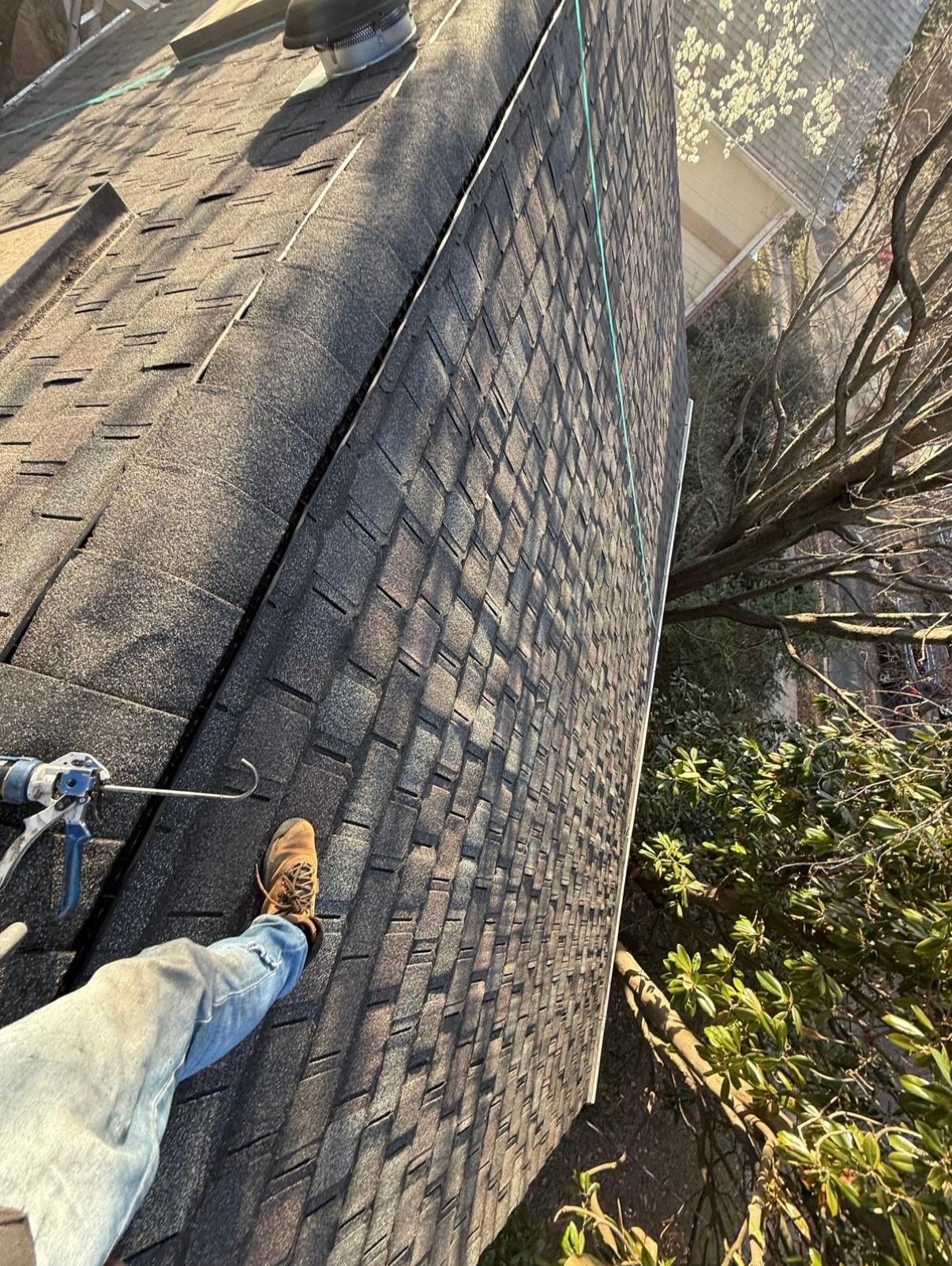 Roof shingles being repaired, with a worker’s legs visible on a steep rooftop near trees.