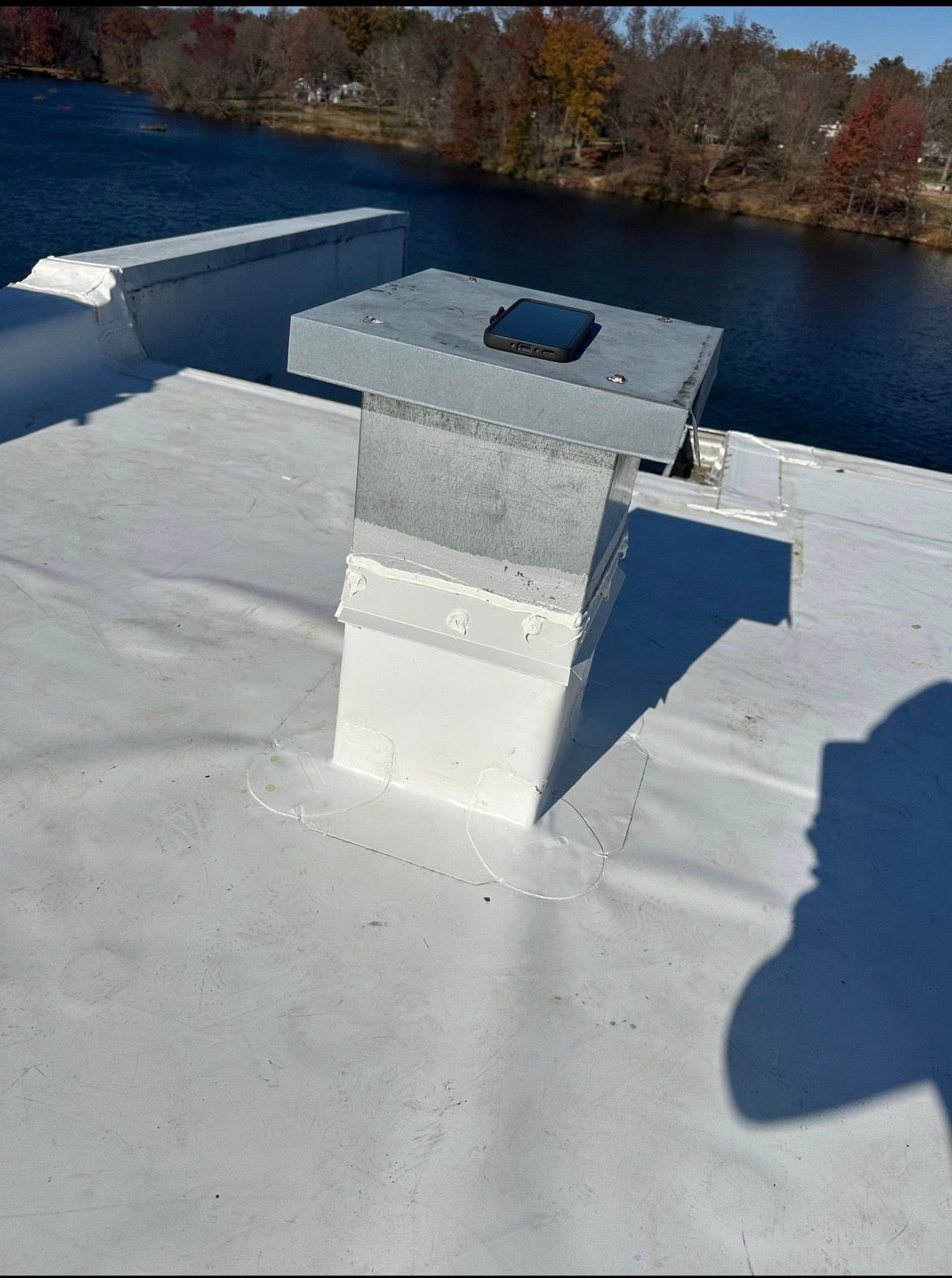 Metal rooftop vent on a white roof beside a river, with autumn trees in the background.