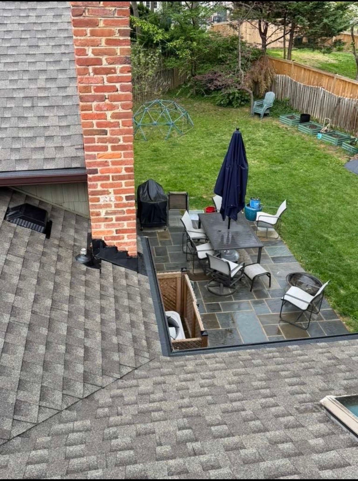 Backyard patio with table, chairs, closed umbrella, and grill beside a brick house and grassy yard