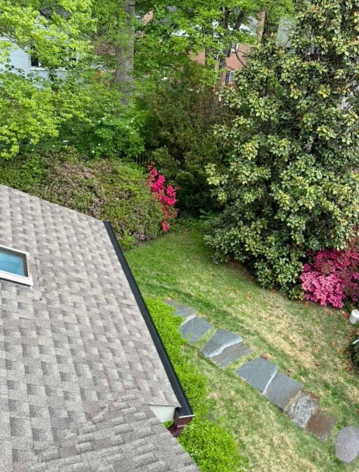 Backyard garden beside a shingled roof, with stepping stones, grass, and pink flowering shrubs.
