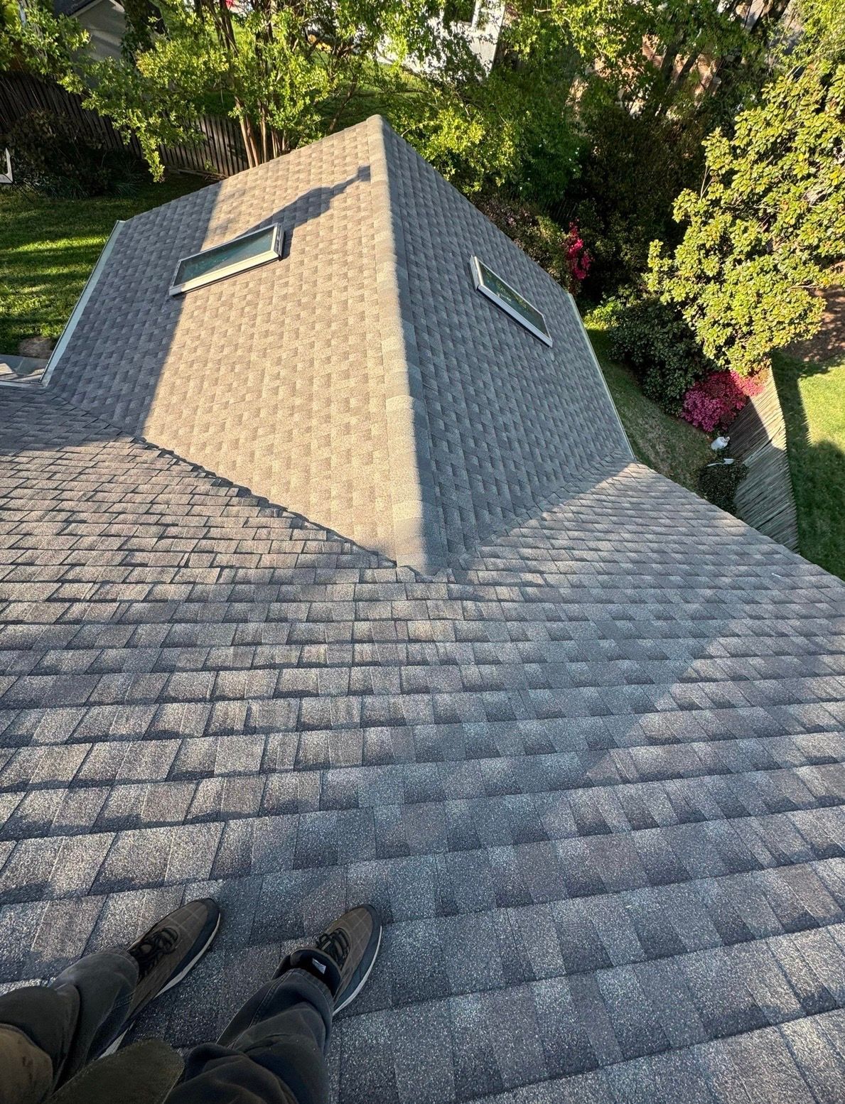 View from a rooftop shingles with feet visible, overlooking a triangular roof peak and green trees below
